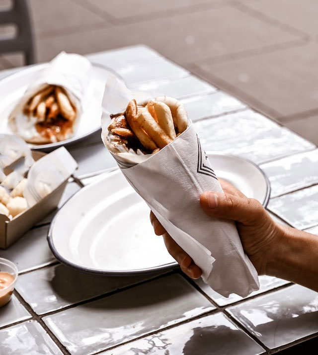 Hand clutching a Greek pita wrapped in a napkin with a table in the background.